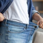 A person in a white T-shirt and unbuttoned denim shirt holds out the waistband of their loose blue jeans, showing they have lost weight.