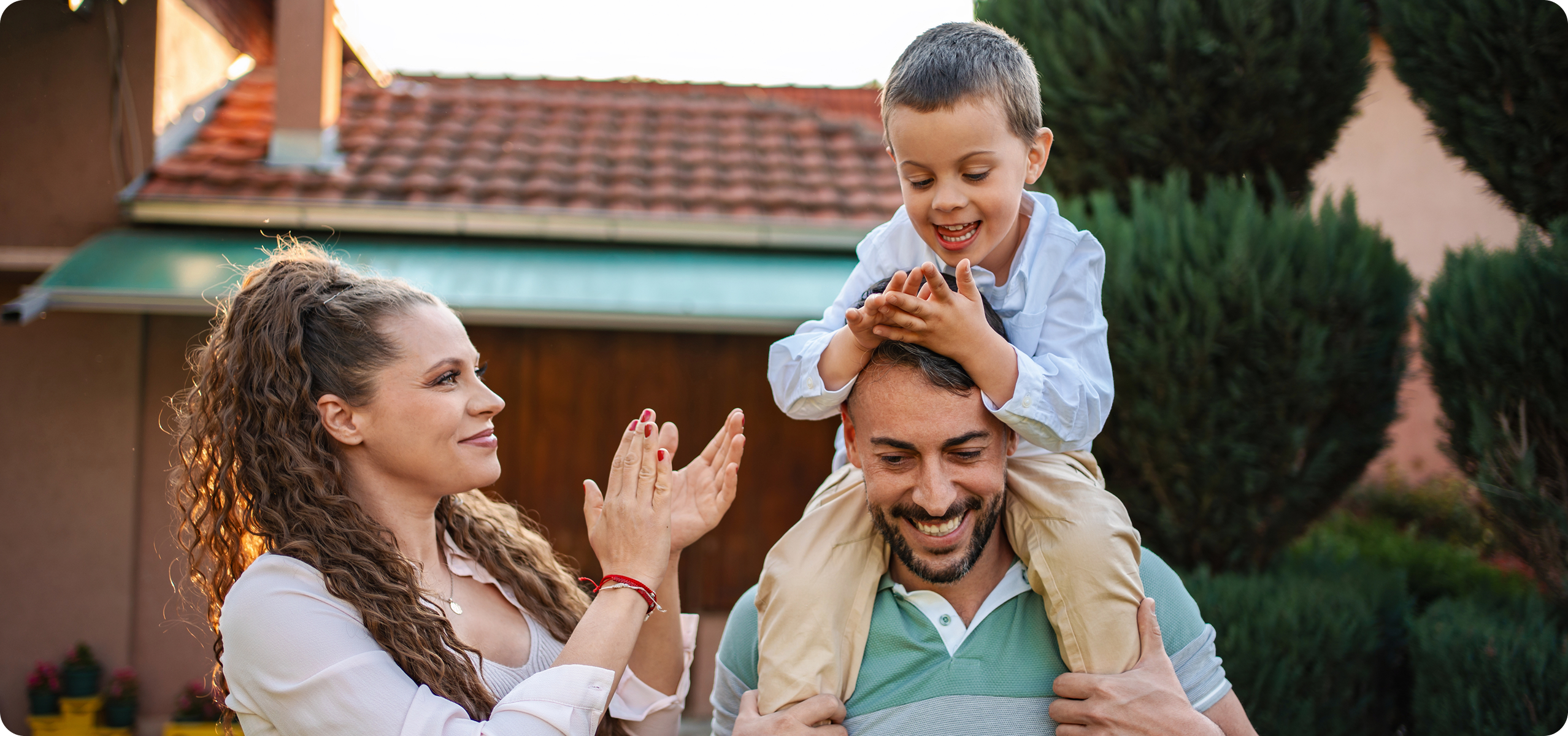 A smiling man carries a happy young boy on his shoulders whilst a woman beside them claps and smiles, outside in front of a house with trees and a tiled roof—a joyful moment celebrating Men’s Health and family togetherness.