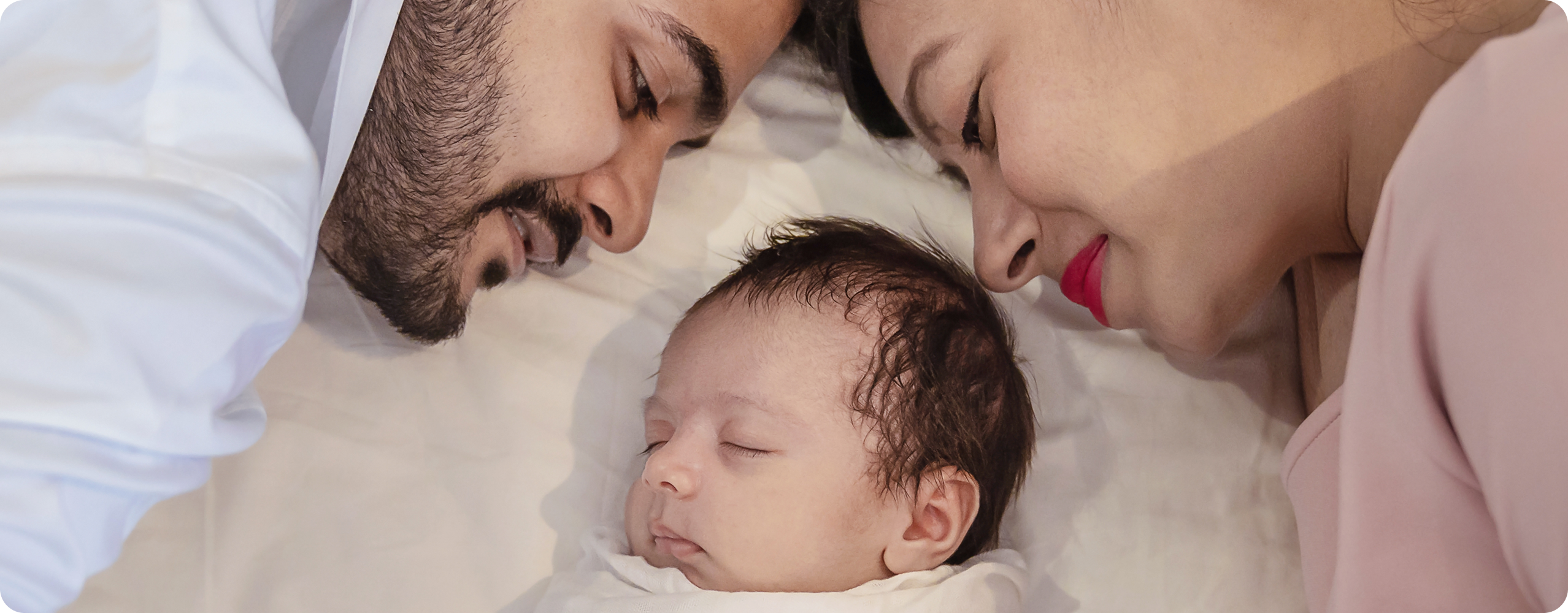 A man and woman lie on either side of a sleeping baby, all facing inwards and smiling gently after the baby's recent circumcision, creating a warm and loving family moment on a white bed.
