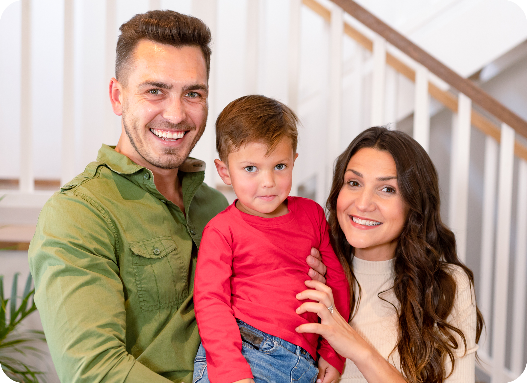 A smiling man and woman stand indoors in front of stairs, holding a young boy in a red shirt. Highlighting Men’s Health, the group pose closely together and look at the camera, radiating happiness and well-being.