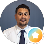 A man with short curly hair, a goatee, and a moustache wears a white shirt and dark tie, posing in front of a blue background. A round badge with a star in the lower right highlights Specialist Men’s Surgical Care.