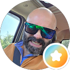 A bearded man wearing dark sunglasses and a brown jacket sits in a car and smiles. Sunlight shines through the window. A colourful star badge icon, highlighting Specialist Men’s Surgical Care, appears in the bottom right corner of the image.