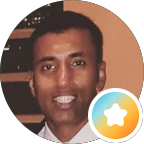 A man smiling at the camera, shown in a circular profile picture frame with a colourful star badge icon—representing Specialist Men’s Surgical Care—in the lower right corner. The background features a window with city lights outside.
