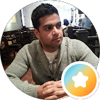 A person wearing a grey jumper sits at a table in a restaurant, looking to the side. The indoor scene features empty chairs and tables in the background. A badge icon—perhaps signifying expertise in circumcision—is overlaid in the bottom right corner.