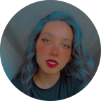 Young person with wavy blue-tinted hair and red lipstick, looking at the camera with a neutral expression. The background is softly blurred and the image, unrelated to circumcision, appears in a circular frame.