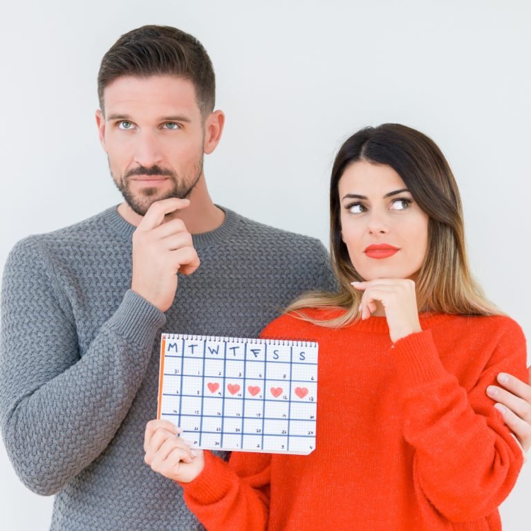 A man and woman stand together, both looking thoughtful. The woman holds a calendar marked with hearts on several days. The man has his hand on his chin, and the woman points at the calendar whilst glancing sideways.