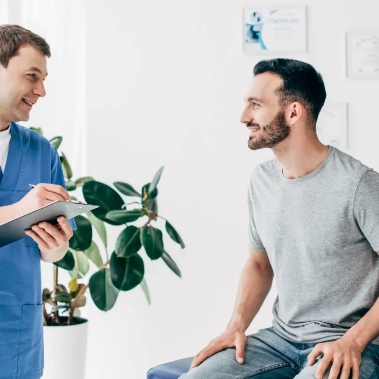 A doctor in blue scrubs talks with a seated male patient during a consultation in a bright clinic at Gentle Procedures UK