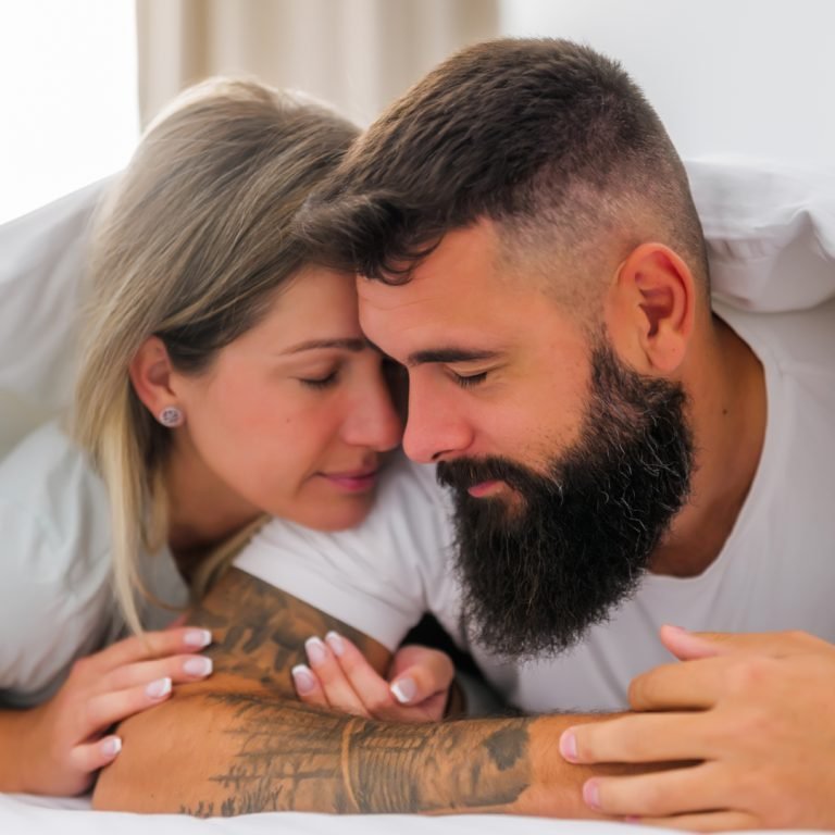 A couple lies close together under a white duvet, facing each other with eyes closed and peaceful expressions, suggesting intimacy and affection.
