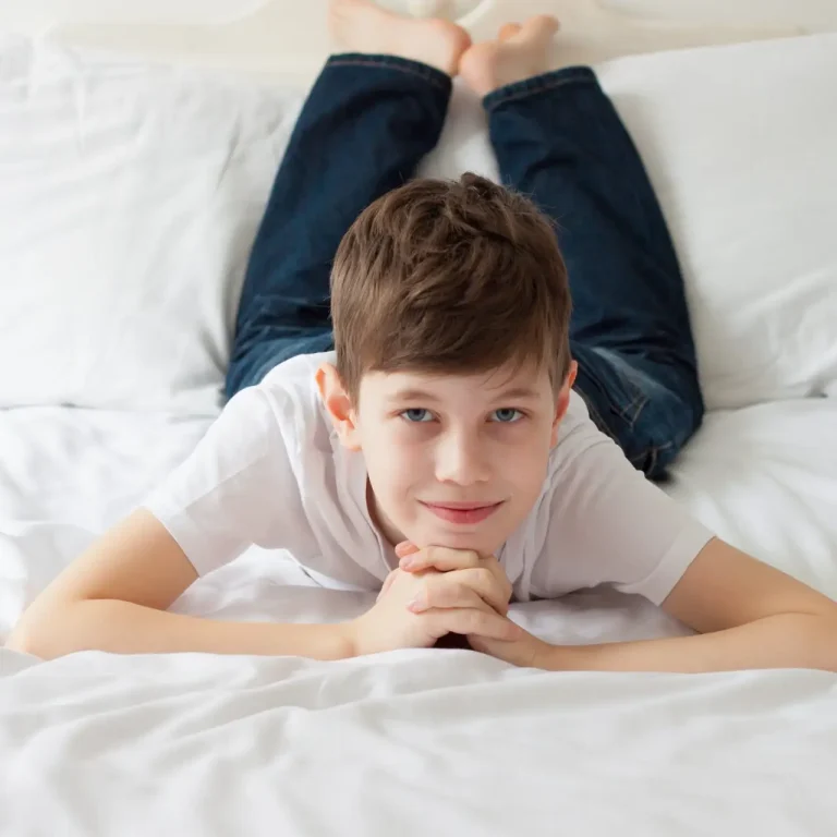 A boy with short brown hair, wearing a white T-shirt and jeans, lies on his stomach on a white bed with his hands clasped under his chin, smiling softly at the camera.