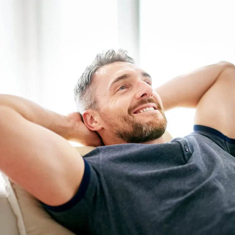 A man with short hair and a beard is smiling and relaxing on a sofa with his hands behind his head, wearing a blue shirt, in a bright room, reflecting the comfort and confidence after visiting a men's health clinic Birmingham.