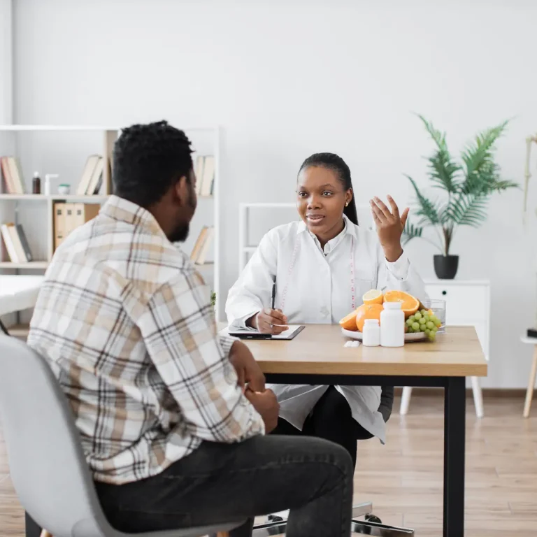A doctor in a white coat sits at a desk, discussing weight loss for men in Birmingham with a patient. There are fruit, a clipboard, and medicine bottles on the desk. Shelves and a skeleton model are in the background.