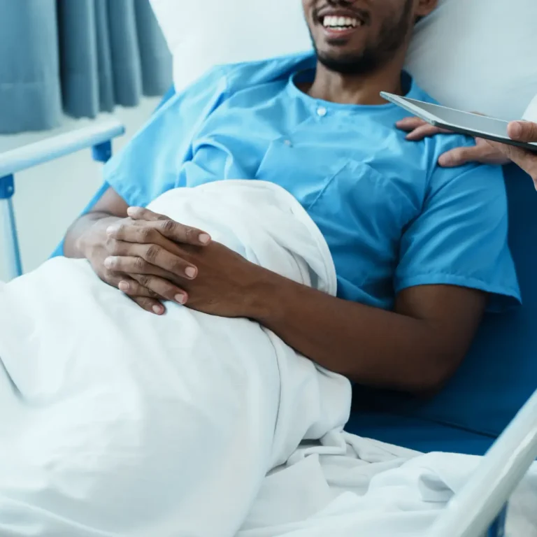 A patient in a hospital bed, smiling and wearing a blue gown, has hands resting on a white blanket. A medical professional stands beside the bed, holding a clipboard or tablet.