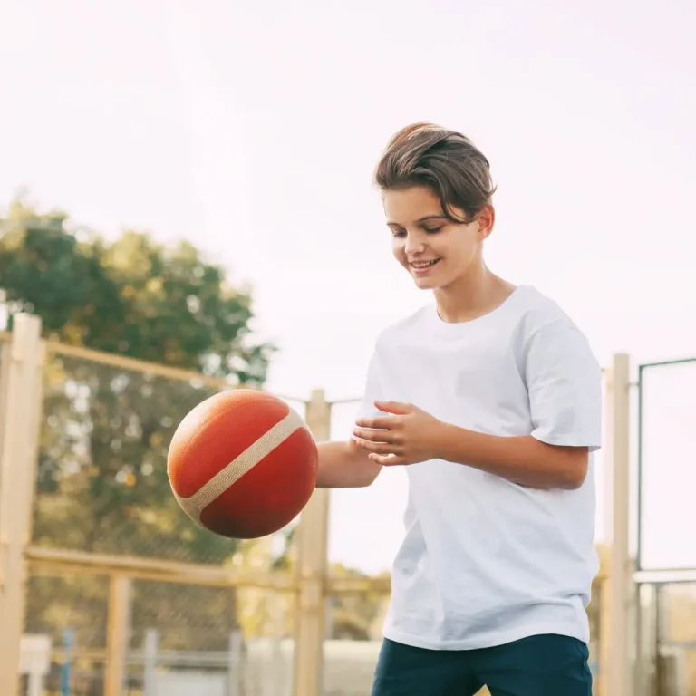 A smiling teenage boy in a white T-shirt dribbles a basketball on an outdoor court with a fence and trees in the background.