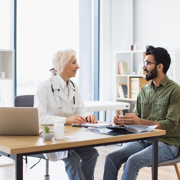 A doctor and a patient are sitting opposite each other at a desk in a bright medical surgery, discussing X-ray images. The doctor is smiling and holding a pen, while the patient listens attentively.