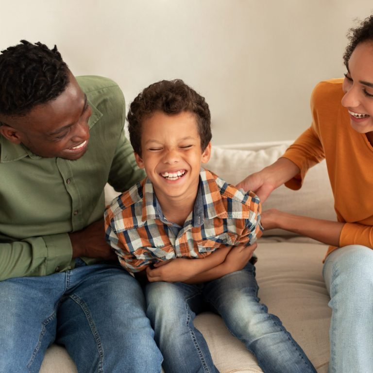 A smiling boy sits on a sofa between two adults, all laughing together. The adults appear to be playfully tickling the boy, creating a joyful and playful family moment.