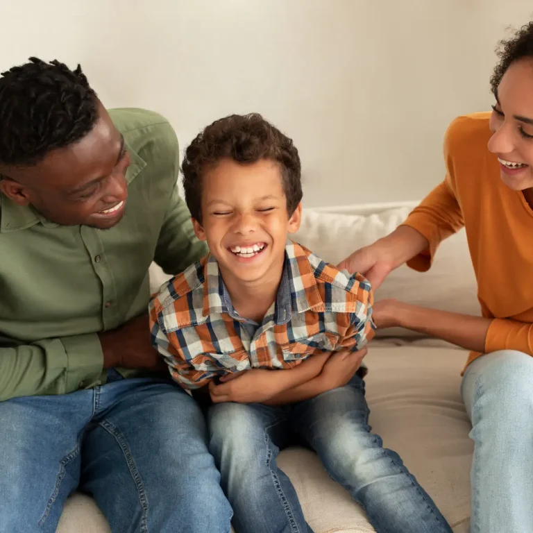 A smiling boy sits on a sofa between two adults, all laughing together. The adults appear to be playfully tickling the boy, creating a joyful and playful family moment.