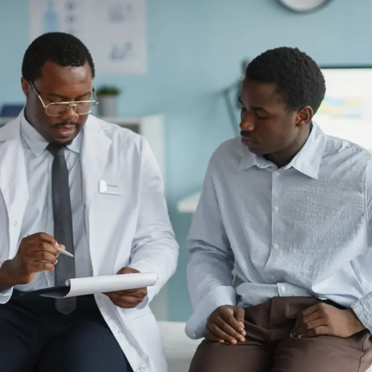 A doctor in a white coat sits beside a patient, reviewing notes on a clipboard. The patient listens attentively. They are in a surgery with medical charts and equipment visible in the background.
