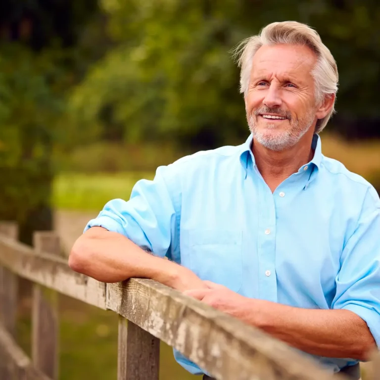 A smiling older man with grey hair and a beard leans on a wooden fence outdoors, wearing a light blue shirt. He embodies the vitality made possible by Specialist Men's Surgical Care, with green trees and grass in the background.
