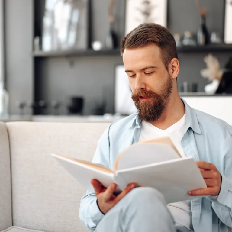 A bearded man sitting on a light-coloured sofa, wearing a light blue shirt and white T-shirt, reads a large book in a modern, softly lit living room.